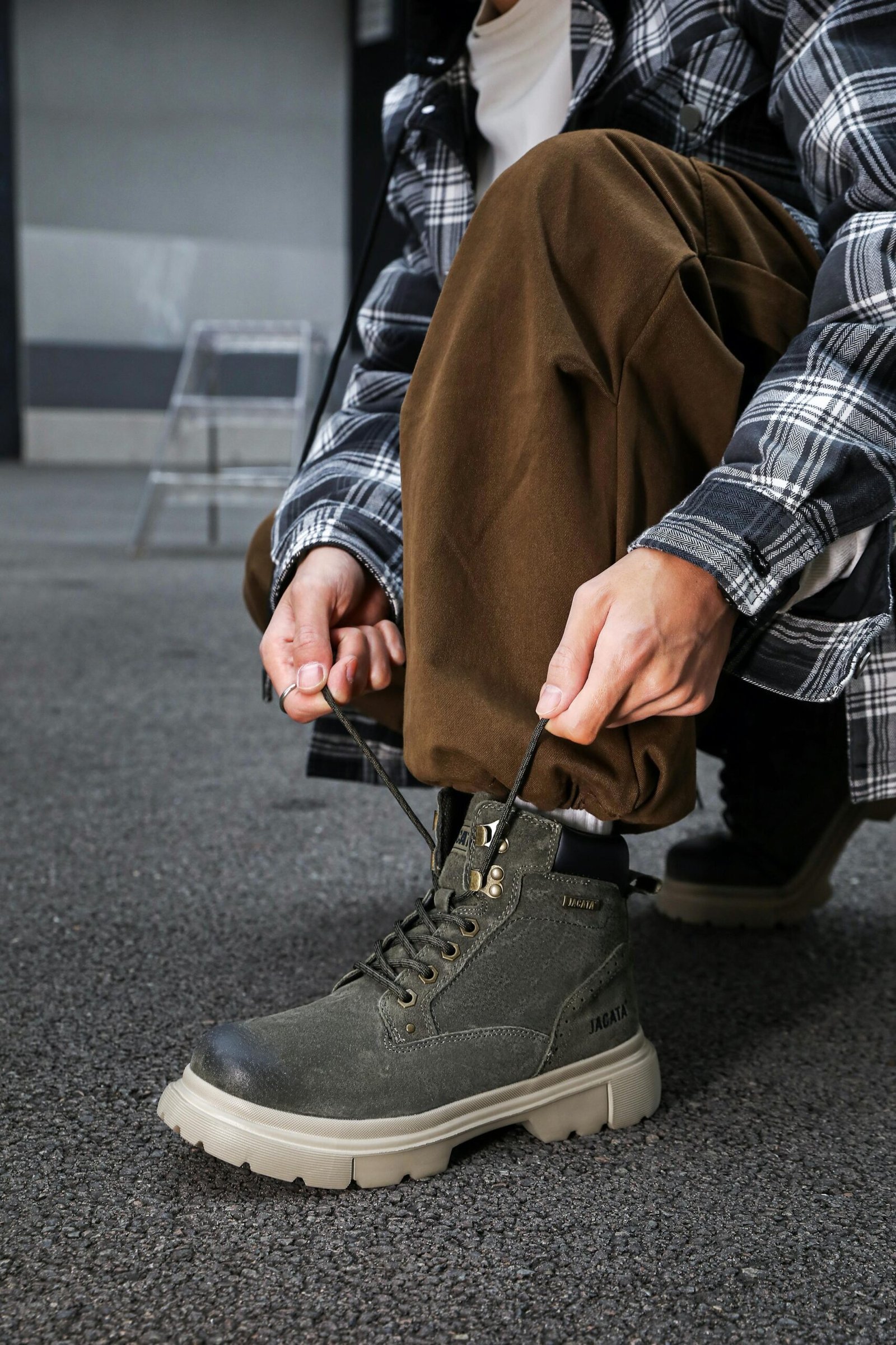 A man in a plaid shirt and brown pants tying the laces of green hiking boots with a thick sole